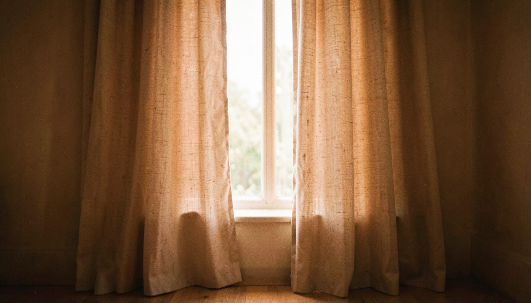 A softly lit interior window dressed with flowing floor-length curtains in a warm neutral linen tone, framing a view of soft natural daylight